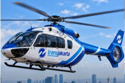 Blue and white Transjakarta helicopter in flight over a city skyline.