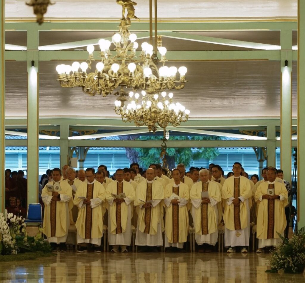 Line of priests in cream and gold vestments standing in a church, holding hymnals beneath a large chandelier during a ceremony
