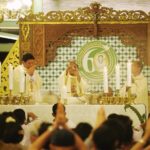 Priests in white robes perform a Catholic mass at an ornate altar, surrounded by lit candles and a gathered worshippers crowd.