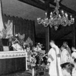 Priests in liturgical robes standing at an altar with candles and flowers during a church ceremony.
