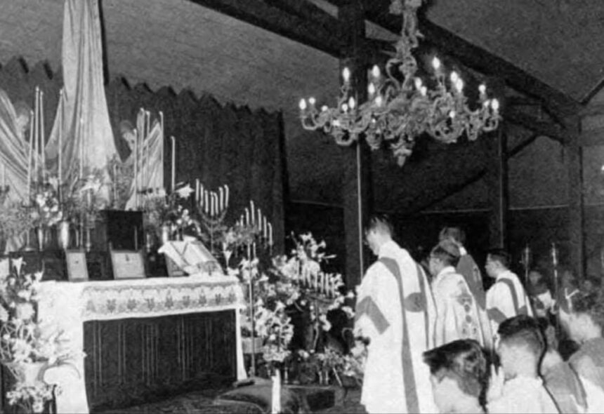 Priests in liturgical robes standing at an altar with candles and flowers during a church ceremony.