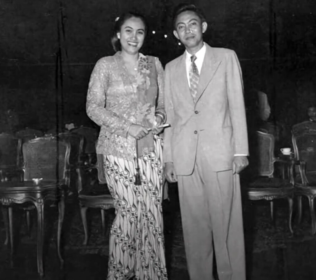 Screenshot_20261009_133039_Instagram - Mabur.co Smiling couple stands together at a formal event; woman in lace top and patterned wide-leg pants, man in a light suit and tie.