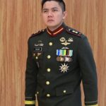 Man in a dark green military dress uniform with gold buttons and many medals, name tag reads TEDDY, standing against a wooden wall.
