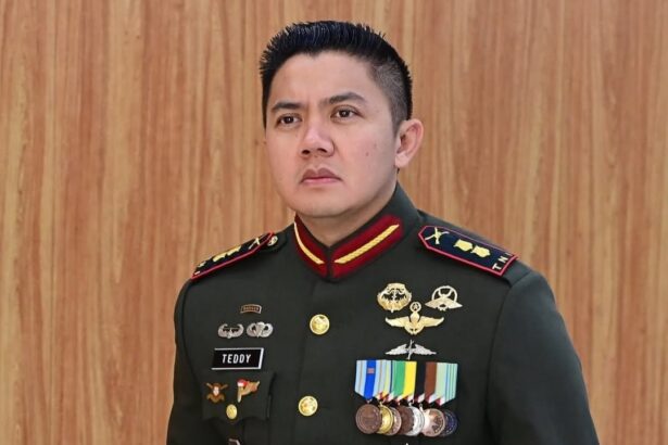 Man in a dark green military dress uniform with gold buttons and many medals, name tag reads TEDDY, standing against a wooden wall.