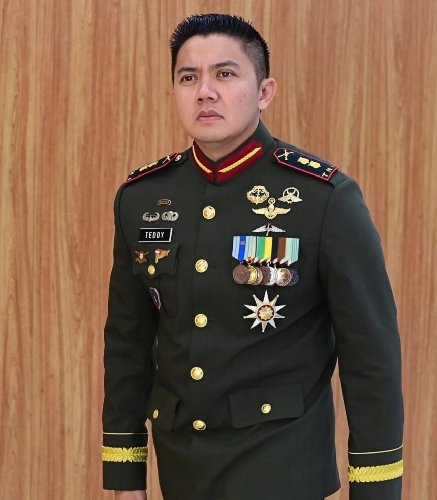 Man in a dark green military dress uniform with gold buttons and many medals, name tag reads TEDDY, standing against a wooden wall.