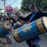 Performer in traditional clothing drums a large blue ceremonial drum in a street parade with several drummers in the background.