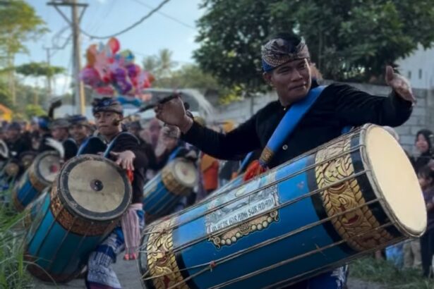 Performer in traditional clothing drums a large blue ceremonial drum in a street parade with several drummers in the background.
