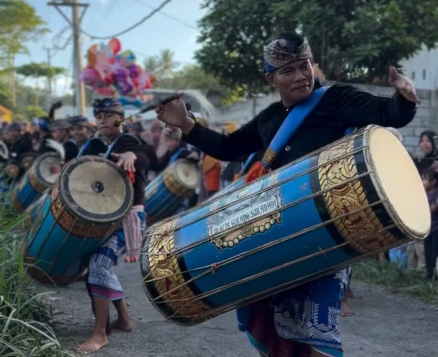 Performer in traditional clothing drums a large blue ceremonial drum in a street parade with several drummers in the background.