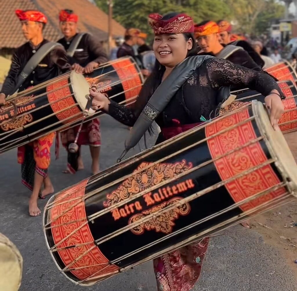 Group of performers in traditional dress playing large decorated drums in a street parade; smiling woman leads the rhythm.