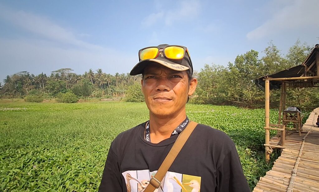 Screenshot_20261010_170640_Gallery - Mabur.co Man in a black shirt with a beige cap and orange sunglasses perched on top, standing in a lush marsh with a bamboo walkway on the right.