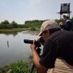 Man with a telephoto lens on a tripod crouches by a calm river while a smiling woman stands on a raised wooden platform, holding a microphone or camera.