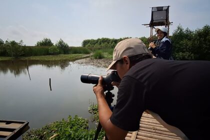 Man with a telephoto lens on a tripod crouches by a calm river while a smiling woman stands on a raised wooden platform, holding a microphone or camera.