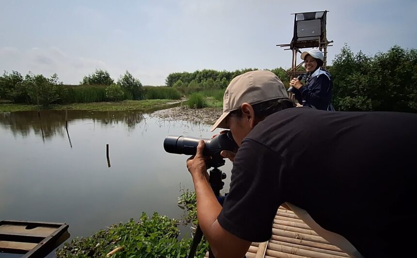 Man with a telephoto lens on a tripod crouches by a calm river while a smiling woman stands on a raised wooden platform, holding a microphone or camera.