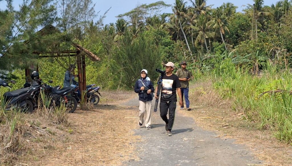 Screenshot_20261010_171251_Gallery - Mabur.co Three people walk along a rural path with camera gear; motorcycles are parked to the left and dense tropical vegetation surrounds them.