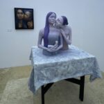 Sculpture of two pale figures with purple hair embracing at a table in a gallery, on a gray patterned cloth.