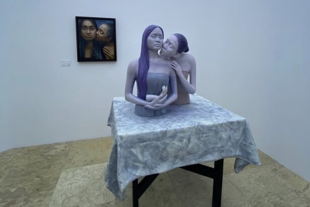 Sculpture of two pale figures with purple hair embracing at a table in a gallery, on a gray patterned cloth.