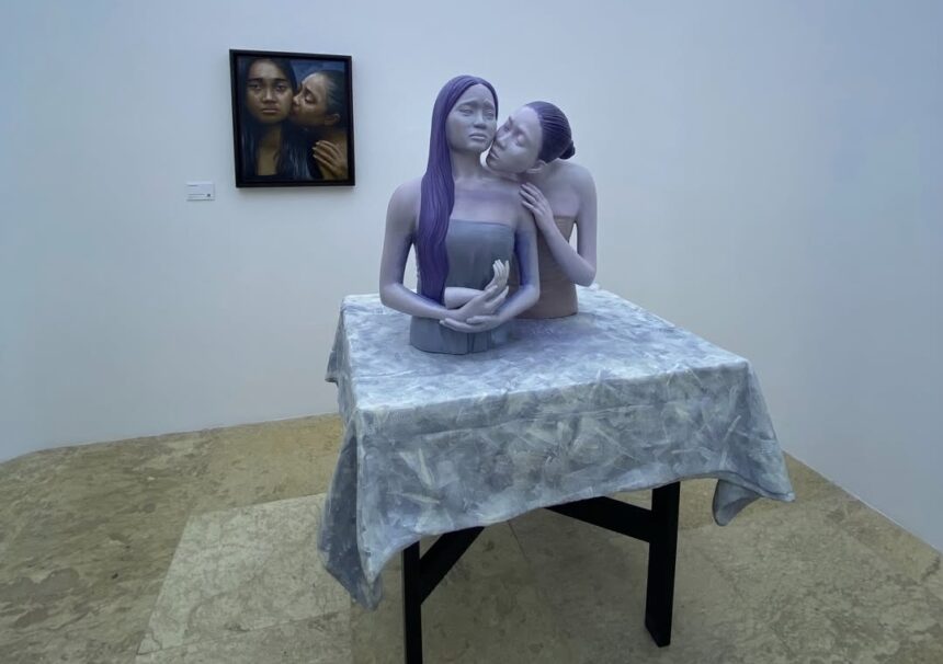 Sculpture of two pale figures with purple hair embracing at a table in a gallery, on a gray patterned cloth.