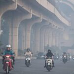 Motorcycles and cars ride on a hazy urban highway beneath a large elevated concrete overpass or viaduct, daytime traffic ahead.