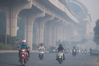 Motorcycles and cars ride on a hazy urban highway beneath a large elevated concrete overpass or viaduct, daytime traffic ahead.