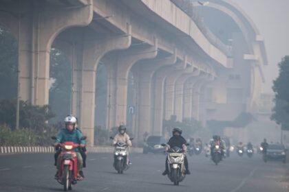 Motorcycles and cars ride on a hazy urban highway beneath a large elevated concrete overpass or viaduct, daytime traffic ahead.