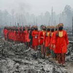 Line of people in bright red traditional attire walking through a smoky, burned landscape after a forest fire, wearing masks.