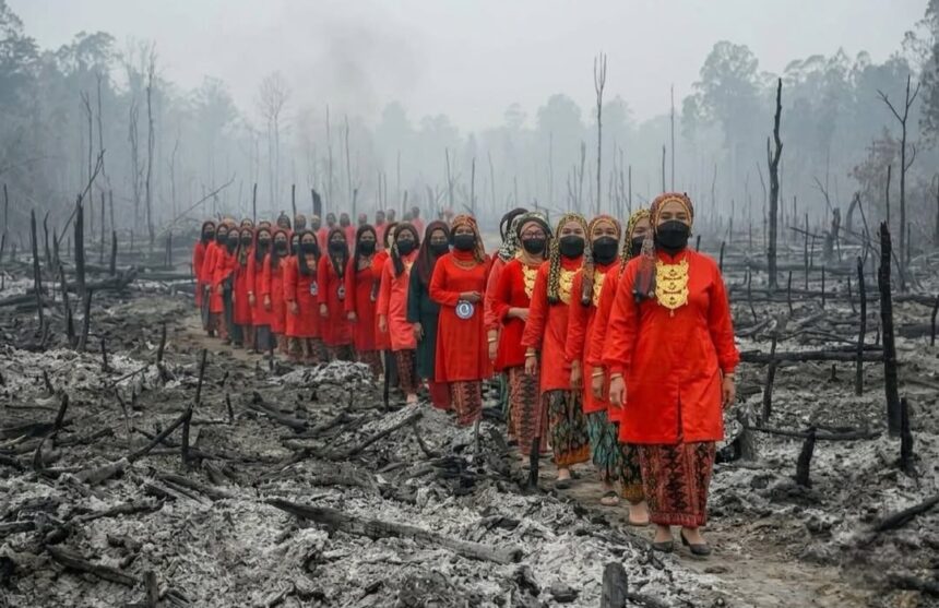 Line of people in bright red traditional attire walking through a smoky, burned landscape after a forest fire, wearing masks.