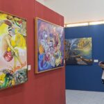 Two visitors stand side by side in an art gallery, admiring large colorful paintings on red and blue walls.