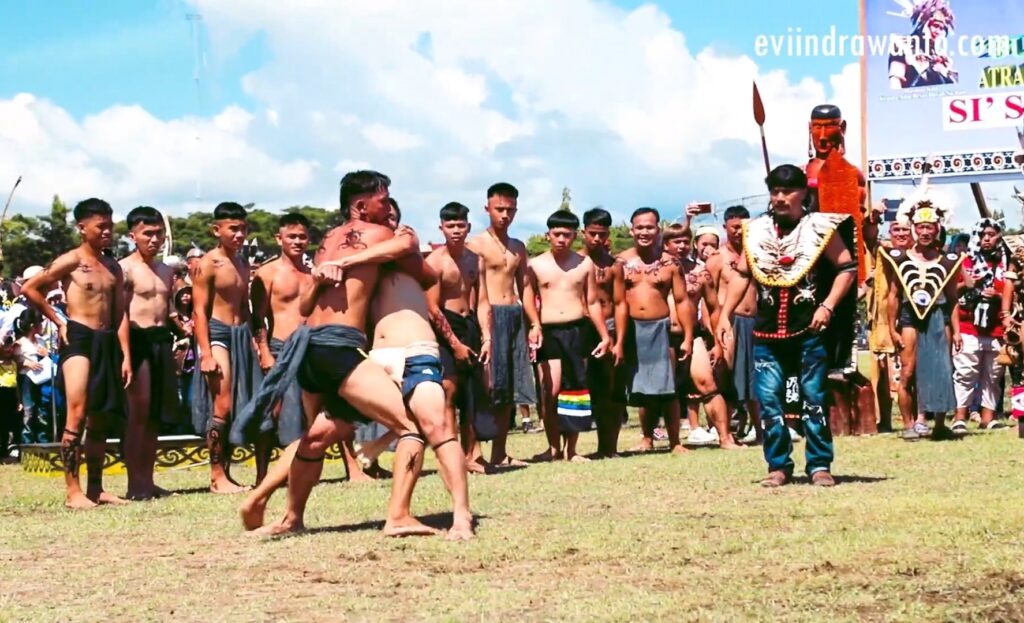 Group of shirtless men in traditional loincloths watching a ceremonial wrestling match outdoors; two competitors grapple on a grassy field while spectators, including a man in ornate ceremonial attire, stand nearby under a bright blue sky.
