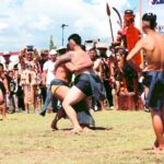 Two shirtless wrestlers grapple in a grassy outdoor festival as a crowd watches, some in traditional attire and costumes in the background.