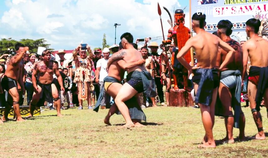 Two shirtless wrestlers grapple in a grassy outdoor festival as a crowd watches, some in traditional attire and costumes in the background.