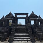 Ancient stone temple gateway with central opening, flanked by two pyramidal pavilions and broad stone stairs under a blue sky.