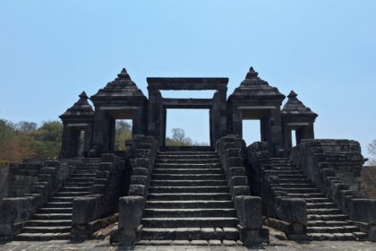 Ancient stone temple gateway with central opening, flanked by two pyramidal pavilions and broad stone stairs under a blue sky.