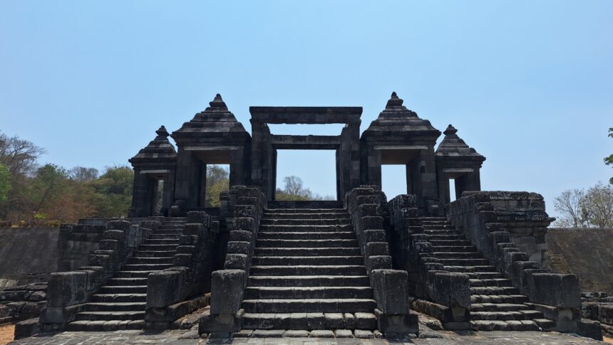Ancient stone temple gateway with central opening, flanked by two pyramidal pavilions and broad stone stairs under a blue sky.