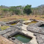 Ancient stone ruin with rectangular limestone basins filled with green water, set in dry grassy landscape and scattered stones.