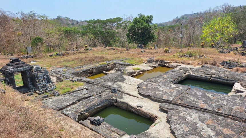 Ancient stone ruin with rectangular limestone basins filled with green water, set in dry grassy landscape and scattered stones.