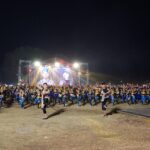 Large group of dancers in blue and black outfits performing on a lit outdoor stage at night, with a big screen and stage lighting in the background.