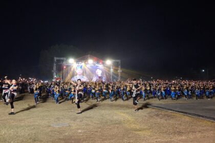 Large group of dancers in blue and black outfits performing on a lit outdoor stage at night, with a big screen and stage lighting in the background.