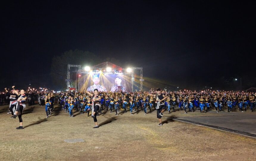 Large group of dancers in blue and black outfits performing on a lit outdoor stage at night, with a big screen and stage lighting in the background.