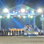 Outdoor nighttime stage ceremony with a metal truss structure and bright multicolored lights; performers in uniforms stand in a long row on the stage
