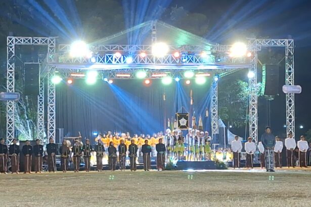 Outdoor nighttime stage ceremony with a metal truss structure and bright multicolored lights; performers in uniforms stand in a long row on the stage