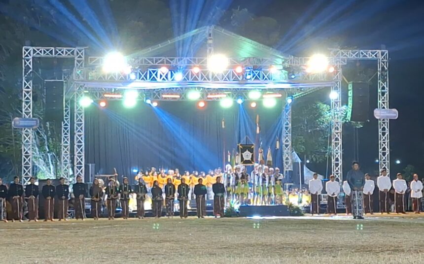 Outdoor nighttime stage ceremony with a metal truss structure and bright multicolored lights; performers in uniforms stand in a long row on the stage