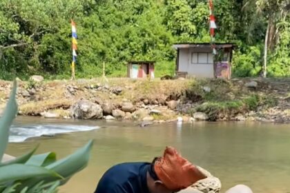 Woman standing in a shallow river, cradling a newborn wrapped in cloth against her chest while standing among rocks and greenery.