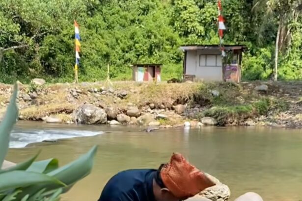 Woman standing in a shallow river, cradling a newborn wrapped in cloth against her chest while standing among rocks and greenery.