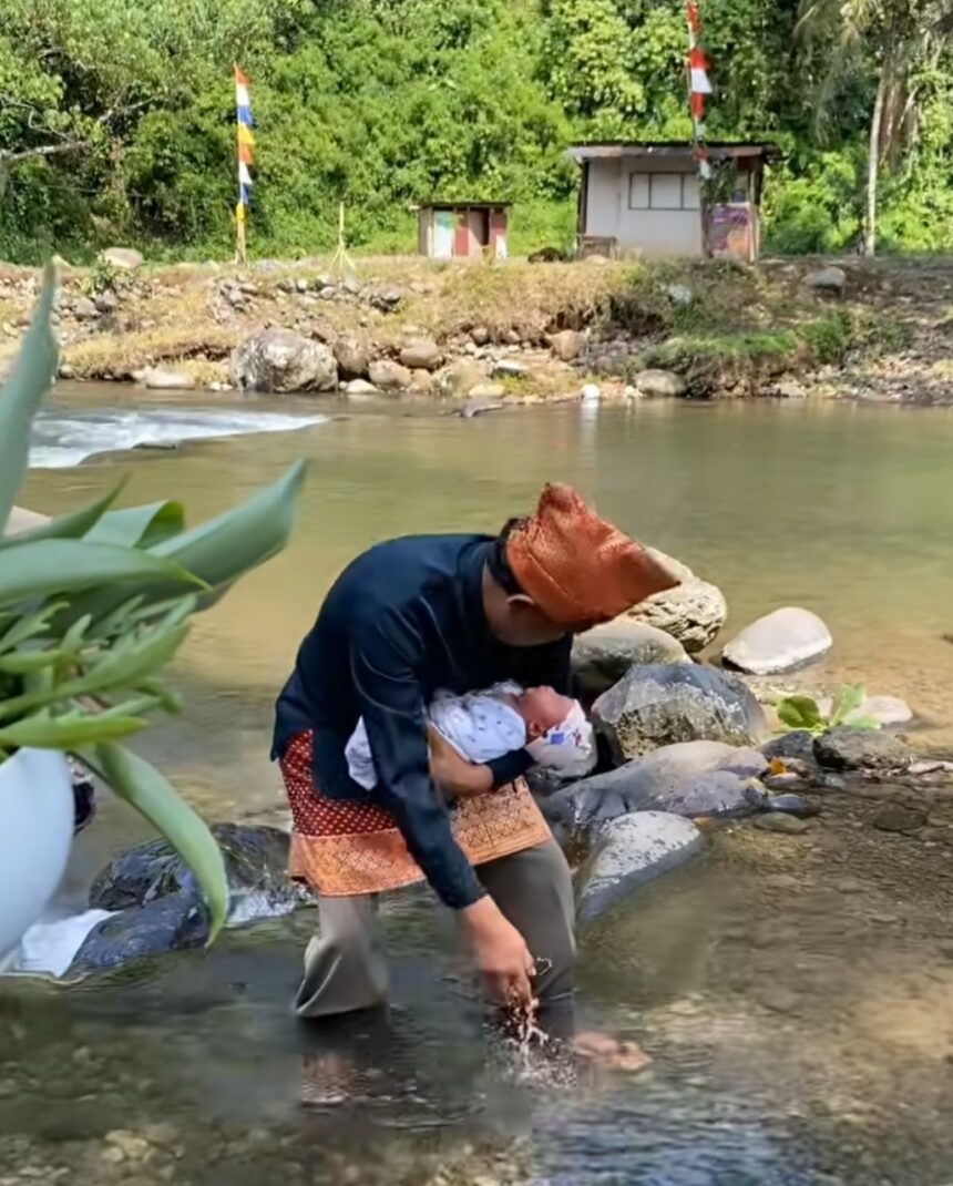 Woman standing in a shallow river, cradling a newborn wrapped in cloth against her chest while standing among rocks and greenery.