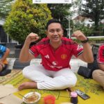 Man in a red Manchester United jersey sits cross‑legged on a yellow patterned mat outdoors, fists raised in celebration, with skewers and bowls of sauce in front of him.