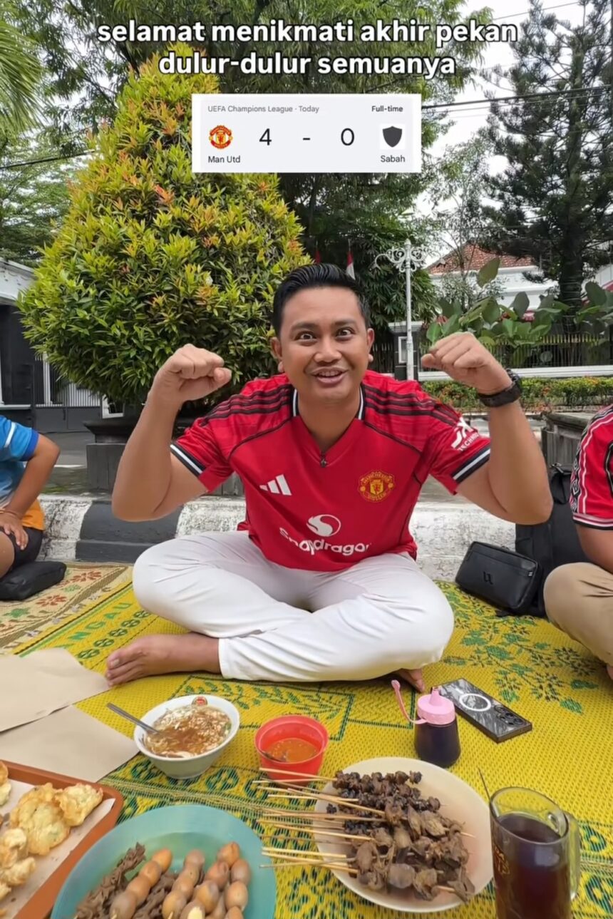 Man in a red Manchester United jersey sits cross‑legged on a yellow patterned mat outdoors, fists raised in celebration, with skewers and bowls of sauce in front of him.