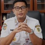 Man in white government uniform with badges sits at desk, hands folded, speaking to camera.