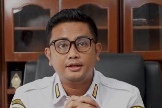 Man in white government uniform with badges sits at desk, hands folded, speaking to camera.