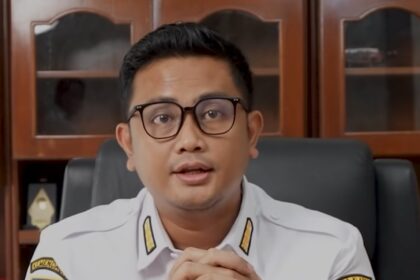 Man in white government uniform with badges sits at desk, hands folded, speaking to camera.