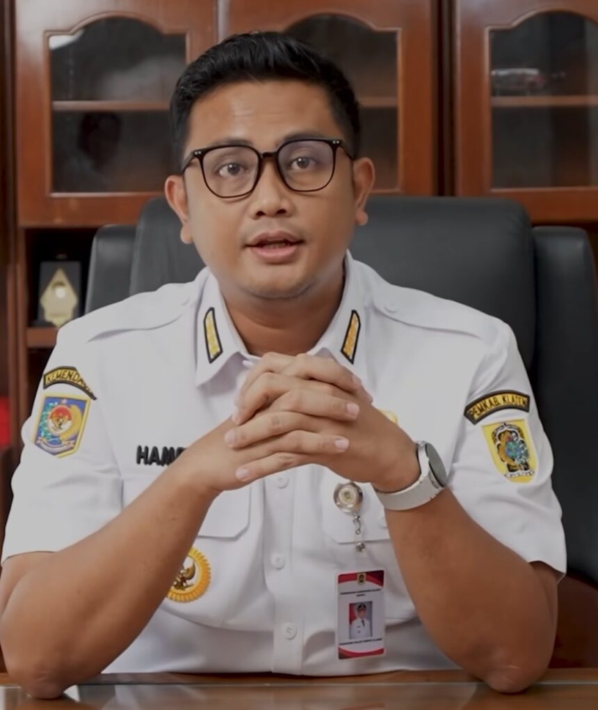 Man in white government uniform with badges sits at desk, hands folded, speaking to camera.